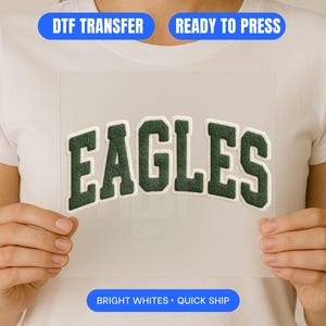 May include: A white t-shirt with a DTF transfer of the word "EAGLES" in a dark green, arched, collegiate-style font. The transfer is ready to press and features the text "DTF TRANSFER", "READY TO PRESS", and "BRIGHT WHITES • QUICK SHIP".