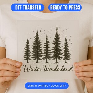 May include: A white DTF transfer featuring a black and white illustration of five pine trees and the words "Winter Wonderland". The transfer is held up by a person wearing a white t-shirt.