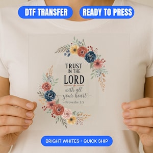 May include: A clear, rectangular DTF transfer featuring the text "TRUST IN THE LORD with all your heart - Proverbs 3:5" surrounded by a floral wreath in shades of red, blue, yellow, and pink. The transfer is held up against a white t-shirt. The words "DTF TRANSFER", "READY TO PRESS", and "BRIGHT WHITES • QUICK SHIP" are also visible.