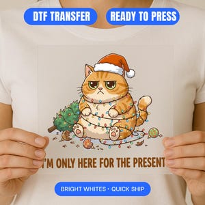 Könnte beinhalten: DTF-Transfer mit einer grimmigen, orangefarbenen Tabby-Katze mit Weihnachtsmütze und Lichterketten. Die Katze befindet sich neben einem kleinen Weihnachtsbaum und Kugeln. Der Text "I'M ONLY HERE FOR THE PRESENT" steht darunter.