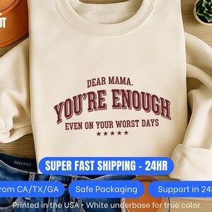 May include: A cream-coloured sweatshirt featuring the burgundy text "DEAR MAMA. YOU'RE ENOUGH EVEN ON YOUR WORST DAYS". The sweatshirt is shown with blue denim jeans. The text is in a bold, collegiate-style font.