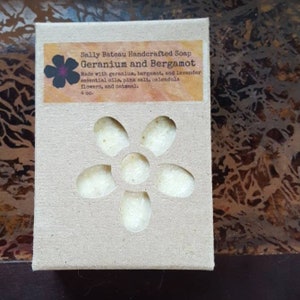 May include: A brown cardboard box with a white label that reads "Sally Bateau Handcrafted Soap Geranium and Bergamot Made with geranium, bergamot, and lavender essential oils, pink salt, calendula flowers, and oatmeal. 4 oz."