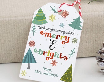 Puede incluir: Etiqueta de regalo blanca con un lazo de cuerda rojo y blanco. La etiqueta tiene una ilustración colorida de árboles de Navidad y copos de nieve. El texto en la etiqueta dice "thank you for making school merry & bright from, Mrs. Johnson".