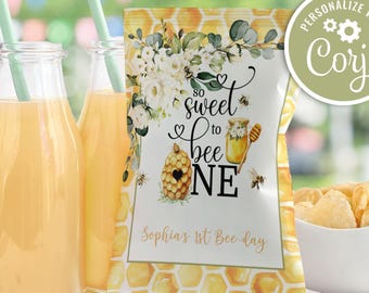 May include: A party scene with two glass bottles of orange juice with green straws, a snack bag with a bee-themed design, and a bowl of chips. The bag reads "So sweet to bee ONE" and "Sophia's 1st Bee-day."