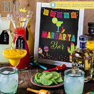 Op de afbeelding: Een krijtbord met de tekst "Margarita Bar" in kleurrijke letters, omringd door feestelijke decoraties. Er staan limoenen, maracas en een fles tequila op een houten tafel.
