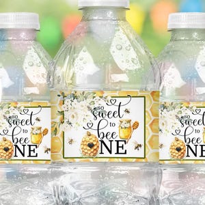 May include: Three clear plastic water bottles with labels that read "Sweet Bee One." The labels feature a honeycomb design, bees, and a honey pot. The words "instant download" are in a circle in the upper left corner. The bottles are on a wooden surface.
