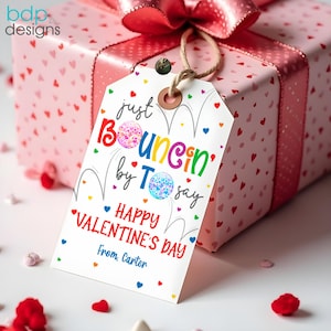 May include: A white gift tag with a colourful design that says "Just Bouncin' by Tsay Happy Valentine's Day" with a heart and the text "From, Carter".