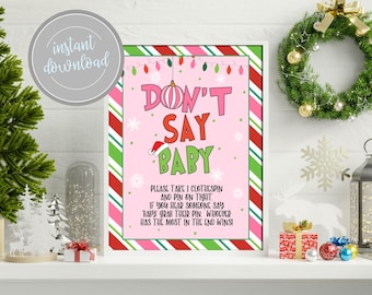 以下が含まれることがあります： 「DON'T SAY BABY」と書かれた、大きなカラフルな文字のピンク色のフレーム入りクリスマスパーティーゲームのサイン。サインには、指示と、サンタの帽子やオーナメントなどのお祝いの装飾が含まれています。背景にはクリスマスリースと木があります。