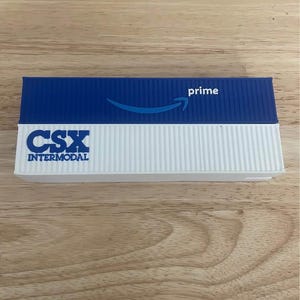 May include: A blue and white CSX Intermodal shipping container model with the word "prime" and an Amazon logo. The model is rectangular and sits on a wooden surface.