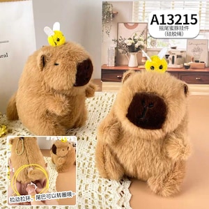 Może przedstawiać: Dwie pluszowe zabawki przedstawiające kapibary z małą pszczołą na głowach. Kapibary są jasnobrązowe z ciemnobrązowymi twarzami. Tekst "A13215" jest widoczny w prawym górnym rogu. Obrazek przedstawia również mniejszy obrazek zabawki kapibary.