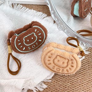 May include: Two plush brown and tan AirPod cases shaped like cookies. The brown case has a bear design with the word "SOF" in a circle. The tan case has a bear design. Both cases have a brown wrist strap.