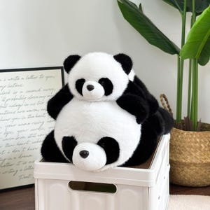 May include: A black and white panda bear soft toy. The panda is resting on a white storage box. The toy has black ears, eyes, and limbs, with a white face and body. The toy appears soft and fluffy.