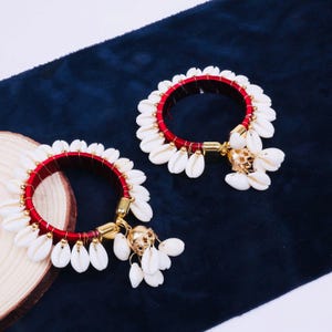 Peut inclure: Deux bracelets rouges et blancs avec des accents dorés. Chaque bracelet est orné de coquillages blancs et de breloques dorées. Les bracelets sont posés sur un fond bleu foncé, créant un contraste saisissant.