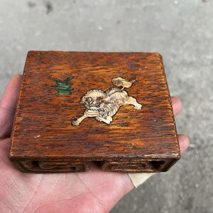 Könnte beinhalten: Kleine, quadratische Holzbox mit eingelegtem Design. Der Deckel zeigt ein geschnitztes, löwenartiges Wesen und eine grüne Pflanze. Das Holz hat eine dunkelbraune Farbe. Die Box wird in einer Hand gehalten.