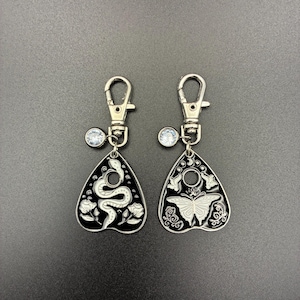 May include: Two black and silver keychains with a planchette design. One features a snake and floral details, the other a butterfly and moon. Each has a silver clasp and a small, clear charm.