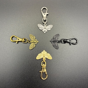 May include: Five metal keychains with moth pendants in silver, gold, and black. The keychains have lobster claw clasps. The moth pendants are detailed with wing and body patterns. The keychains are arranged on a dark gray surface.
