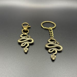 May include: Two bronze-colored snake keychains. One keychain has a clasp, and the other has a chain and a ring. The snakes are coiled with detailed scales and a small, pointed head. The background is a dark gray.