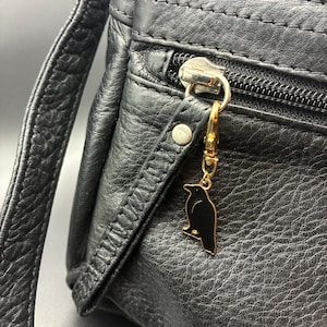 May include: Black leather crossbody bag with a gold-colored zipper and a raven charm. The bag has a textured surface and a long strap. The raven charm is black with gold trim.