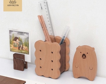 May include: Wooden desk organizer set featuring a biscuit-shaped pen holder, a bear-shaped figurine, and a photo holder. The pen holder contains pencils, a pen, and a ruler. The set is made of light brown wood.