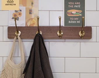 May include: A wooden wall-mounted coat rack with four brass hooks. A beige tote bag and a brown and black patterned coat hang from the hooks. A postcard with the text "YOU ARE THE PATÉ TO MY BÁNH MÌ" is visible.