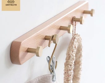 May include: A wooden wall-mounted coat rack with five brass-colored hooks. A metal clip and a beige knitted item hang from the hooks. The rack has the text "HAPPYNESTYN" and a logo in the upper left corner.