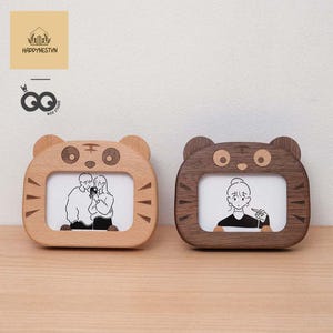 Op de afbeelding: Twee houten fotolijsten in de vorm van tijgers. De ene is lichtbruin, de andere donkerbruin. Elke lijst heeft een rechthoekige opening voor een foto en een cartoonillustratie binnenin. De lijsten hebben oren en tijgerstreepdetails.