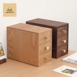 May include: Two wooden desktop organizers, one light and one dark brown, each with three drawers and gold-colored knobs. The light brown organizer is on the left, and the dark brown one is on the right. The logo "HAPPYNESTVN" is in the upper left corner.