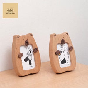Op de afbeelding: Twee houten fotolijsten in de vorm van een beer. De frames zijn lichtbruin met donkerbruine accenten voor de neus en poten. Elk frame bevat een zwart-wit illustratie. De frames staan op een lichtbruin oppervlak.