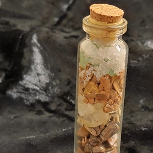 May include: A small, clear glass bottle filled with layers of various natural elements. The bottle is sealed with a cork stopper. The contents include white crystals, green stones, and brown and tan stones.