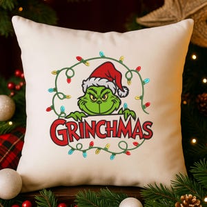 May include: Off-white square pillow featuring a green Grinch character wearing a Santa hat, surrounded by a string of colorful Christmas lights. The word "GRINCHMAS" is written in red below the character.