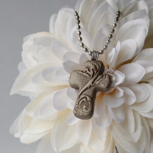 May include: A silver chain necklace with a brown ceramic cross pendant. The cross has a detailed floral design. The necklace is displayed on a white flower.