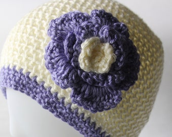 May include: A cream-colored crochet hat with a purple and cream flower accent. The hat has a purple band around the bottom. The hat is displayed on a white mannequin head.