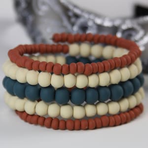 May include: A spiral bracelet composed of small, round beads in alternating colors. The bracelet features bands of cream, teal, and rust-colored beads. The beads are strung together to form a flexible, coiled design, suitable for wrist wear.