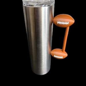 May include: A stainless steel tumbler with a clear lid and a football-shaped handle. The handle is brown with white stitching details, resembling a football. The tumbler is designed to keep beverages cold or hot.