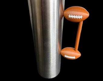 May include: A stainless steel tumbler with a clear lid and a football-shaped handle. The handle is brown with white stitching details, resembling a football. The tumbler is designed to keep beverages cold or hot.