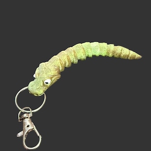 Może przedstawiać: Kapryśny brelok do kluczy przedstawiający segmentowe stworzenie przypominające robaka z głową gada. Stworzenie jest głównie żółto-zielone z teksturowanymi detalami. Jest przymocowany do srebrnego kółka na klucze i karabińczyka.
