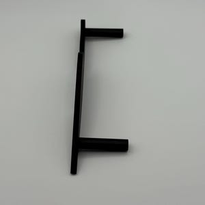 May include: A black, minimalist cabinet pull handle with a T-shaped design. The handle is made of a solid, matte black material and is mounted on a white surface. The handle is designed for easy gripping and pulling.