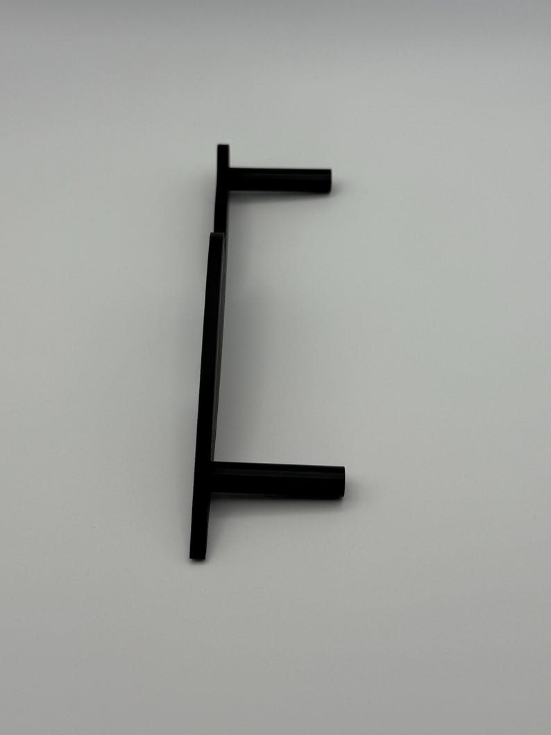 May include: A black, minimalist cabinet pull handle with a T-shaped design. The handle is made of a solid, matte black material and is mounted on a white surface. The handle is designed for easy gripping and pulling.