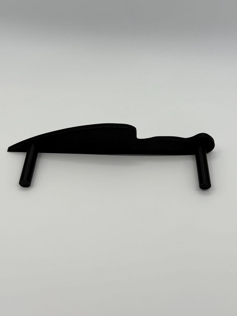 May include: A black knife-shaped object with two vertical supports. The object has a curved blade and a handle-like design. The supports are cylindrical and perpendicular to the blade. The item is likely a display stand.