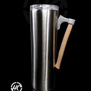 May include: A tall, cylindrical stainless steel tumbler with a clear plastic lid. The handle is shaped like a silver-coloured axe head with intricate embossed detailing, attached to a light tan wooden handle. The logo Hemitts Creations is visible.