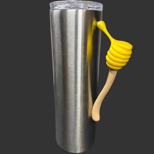 May include: A stainless steel tumbler with a clear lid and a yellow honey dipper handle. The handle is made of a wooden stick and a yellow spiral-shaped dipper. The tumbler is cylindrical and has a brushed metal finish.