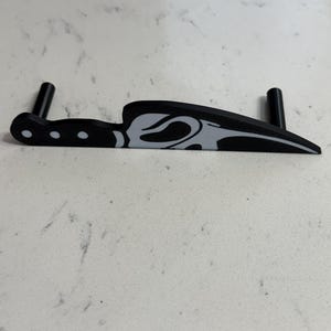 May include: Black knife-shaped object with a gray design on the blade. The handle has three white dots. Two black cylindrical supports are attached to the object, likely for stability. The object is on a white surface.