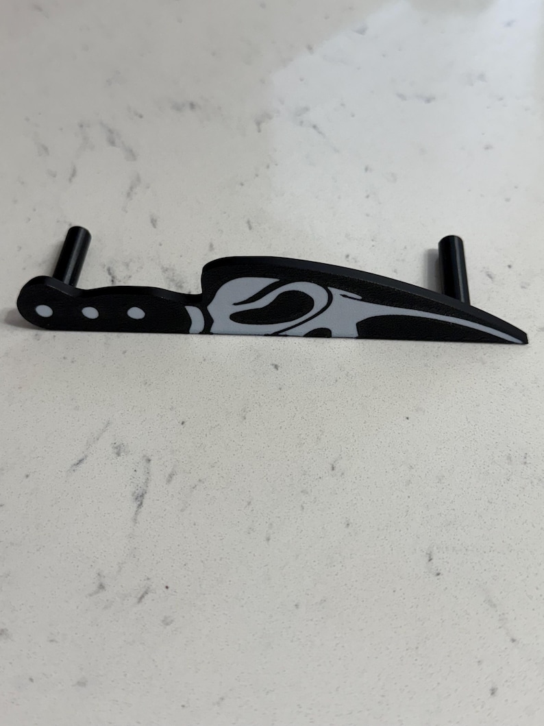 May include: Black knife-shaped object with a gray design on the blade. The handle has three white dots. Two black cylindrical supports are attached to the object, likely for stability. The object is on a white surface.