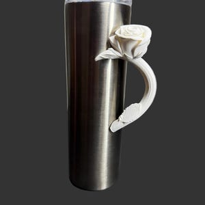 May include: A stainless steel tumbler with a clear lid and a white rose handle. The rose handle is 3D-printed and adds a decorative touch to the insulated drinkware. The tumbler is designed to keep beverages cold or hot.