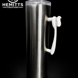 Pode incluir: Um copo cilíndrico alto de aço inoxidável com acabamento em metal escovado e tampa transparente. Uma escova de dentes branca com um pouco de pasta de dentes branca nas cerdas está fixada na lateral como alça. O logótipo HC HEMITTS CREATIONS é visível.