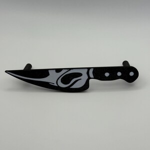 May include: Black and white decorative knife with a stylized design. The knife features a blade with a white design and a black handle with three white dots. The knife is a decorative item.