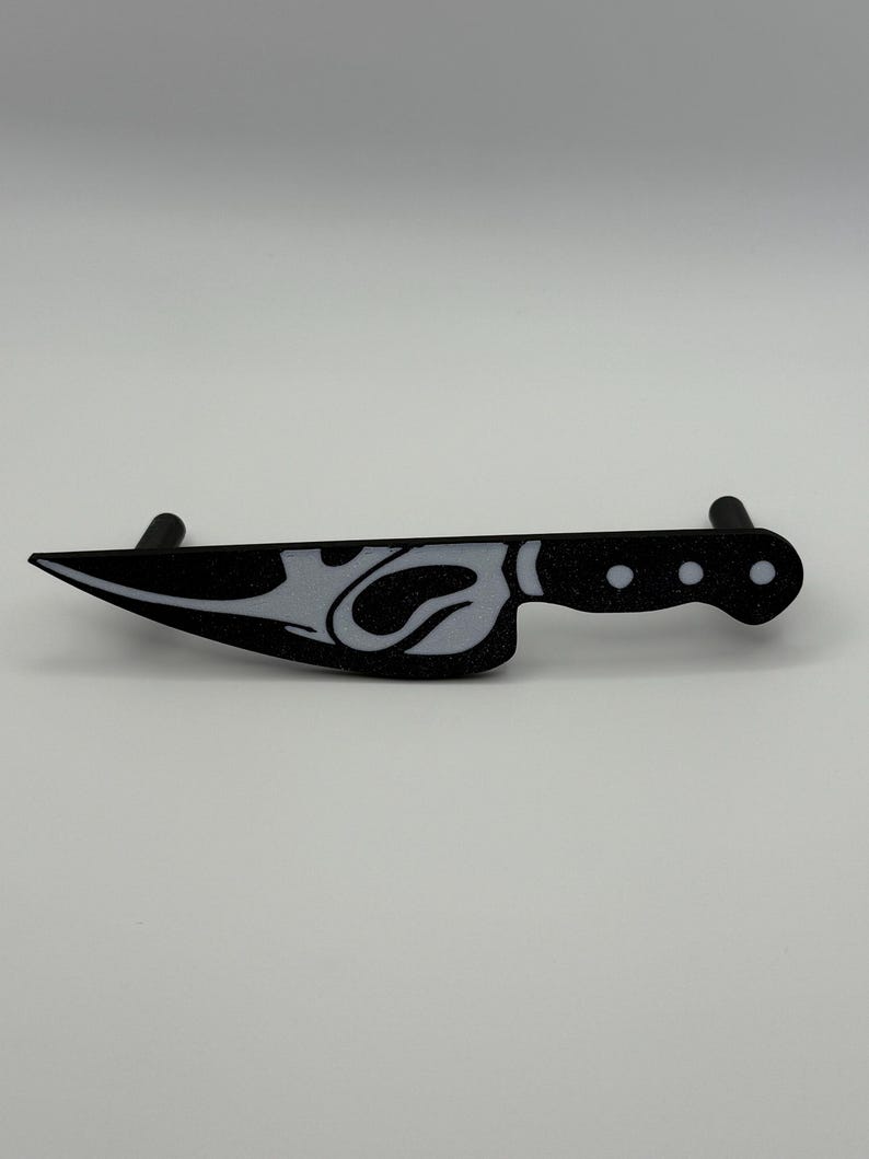May include: Black and white decorative knife with a stylized design. The knife features a blade with a white design and a black handle with three white dots. The knife is a decorative item.