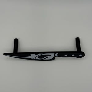 May include: A black and white decorative knife with a stylized flame design. The knife has a handle with three circular indentations and two vertical supports. The knife is made of a solid material and is set against a white background.