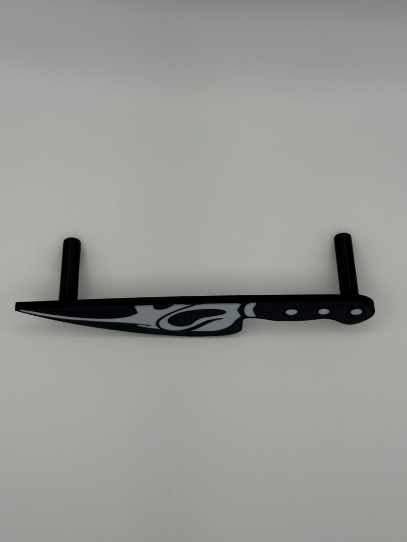 May include: A black and white decorative knife with a stylized flame design. The knife has a handle with three circular indentations and two vertical supports. The knife is made of a solid material and is set against a white background.