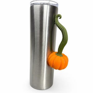 Op de afbeelding: Een hoge, cilindervormige roestvrijstalen beker met een geborstelde metalen afwerking en een doorzichtig deksel. Aan de zijkant van de beker zit een decoratief handvat in de vorm van een feloranje pompoen met een lange, getextureerde olijfgroene steel.